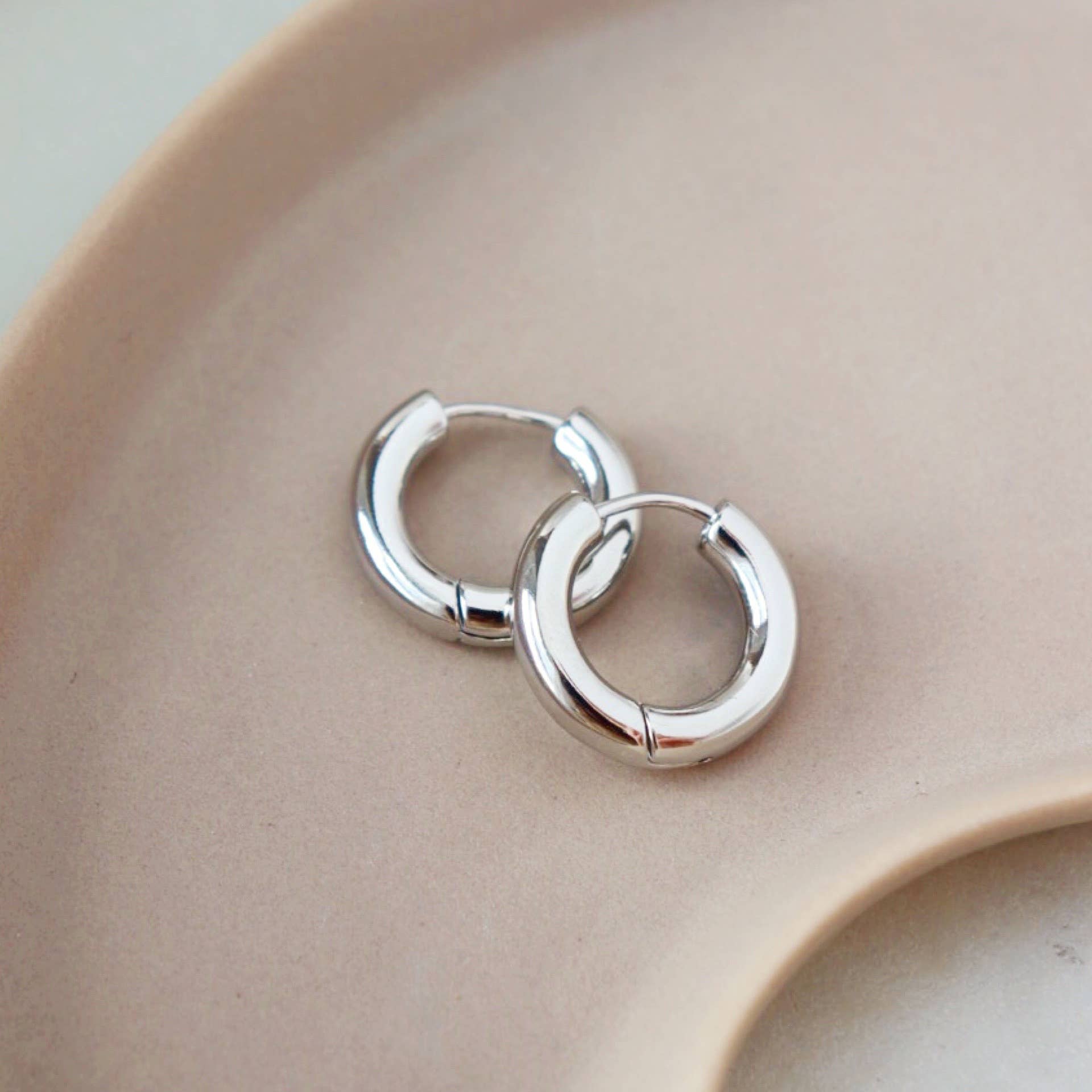 Chunky silver huggie hoop earrings displayed on a soft beige dish
