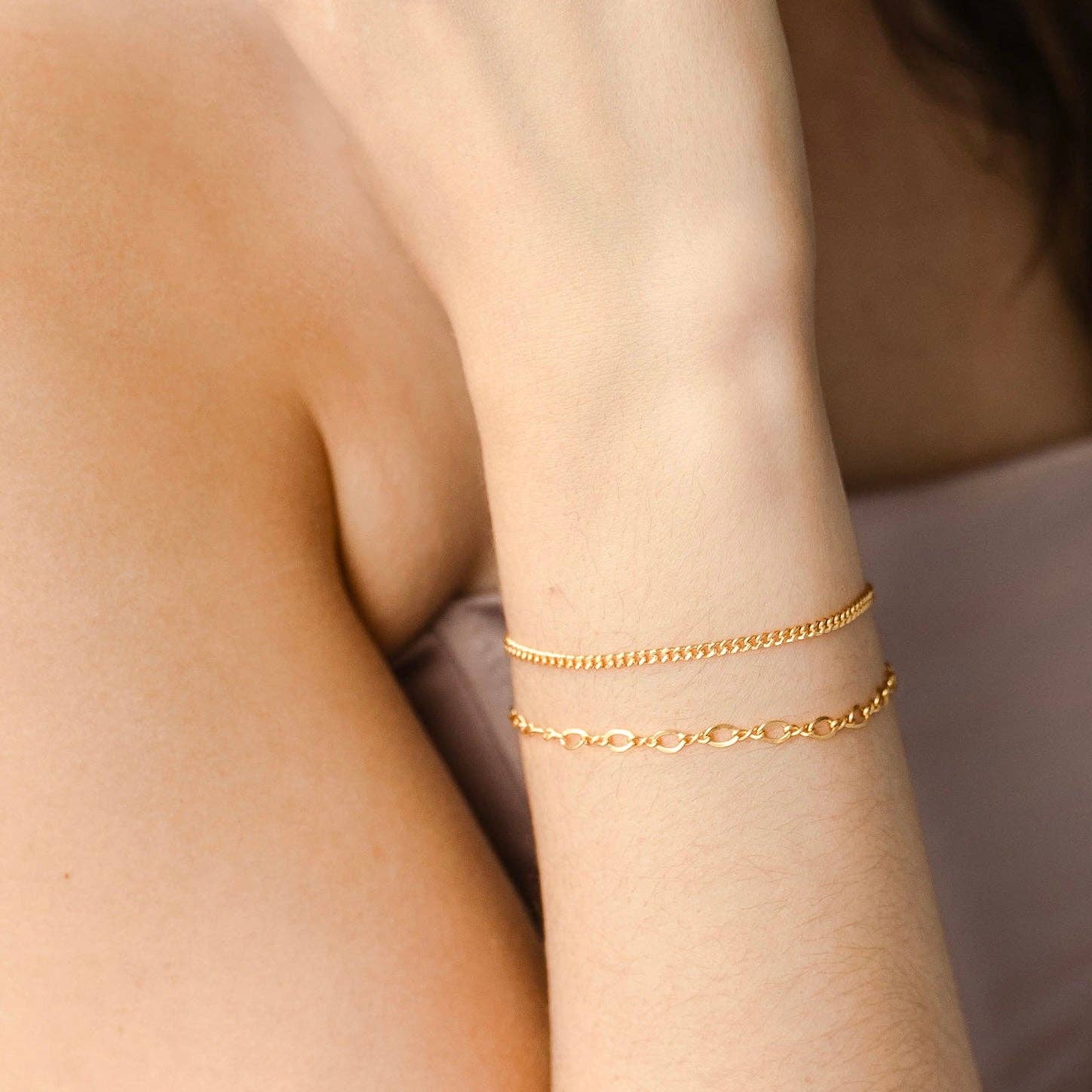 Layered gold chain bracelets, featuring a delicate oval link bracelet and a sleek chain bracelet, worn on a wrist for a stylish and minimal look.
