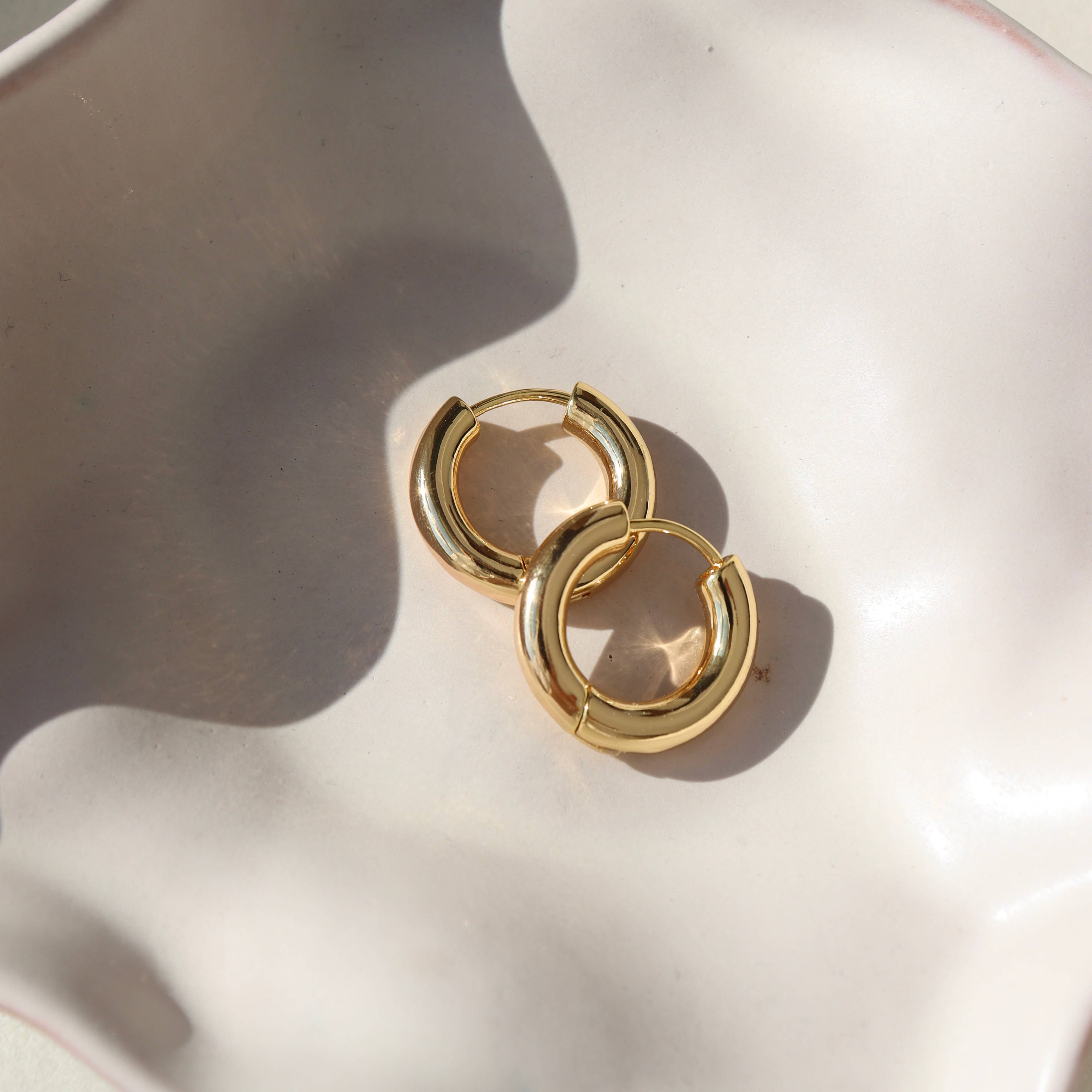 Chunky gold huggie hoop earrings displayed on a soft cream-colored dish