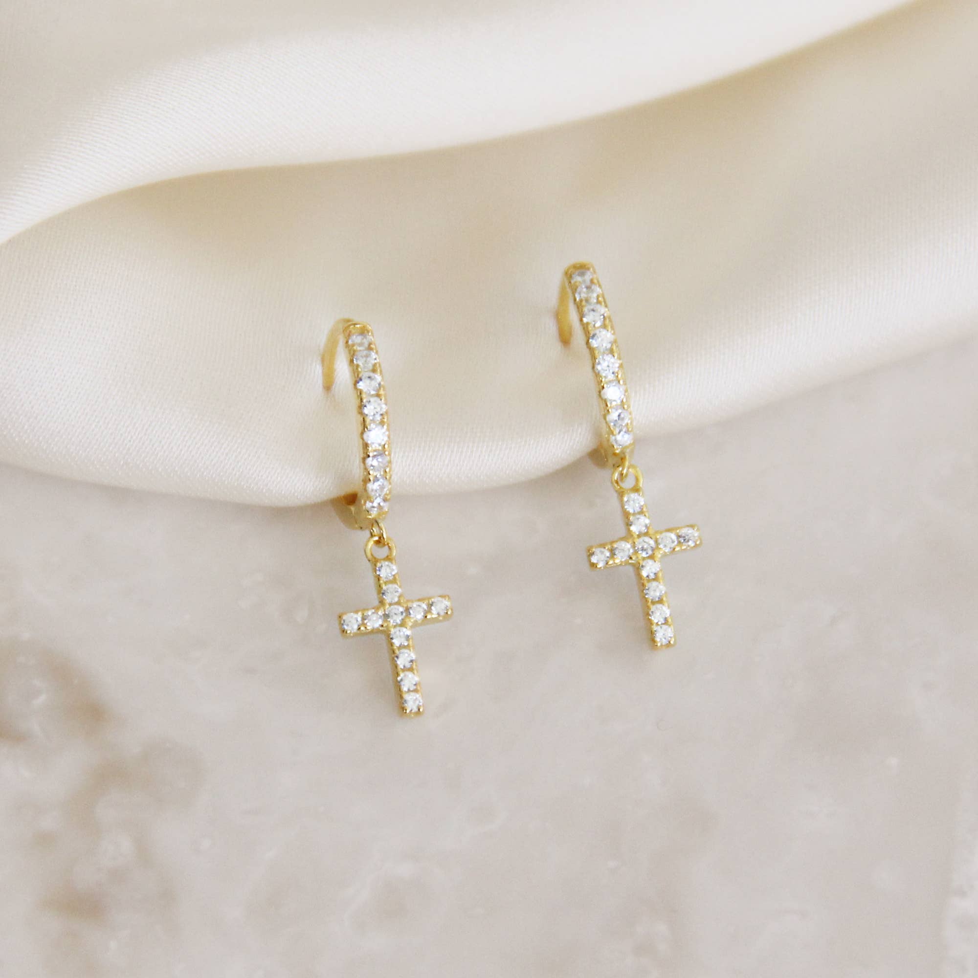 Close-up view of sparkling cross hoop earrings with cubic zirconia stones on a delicate fabric background.