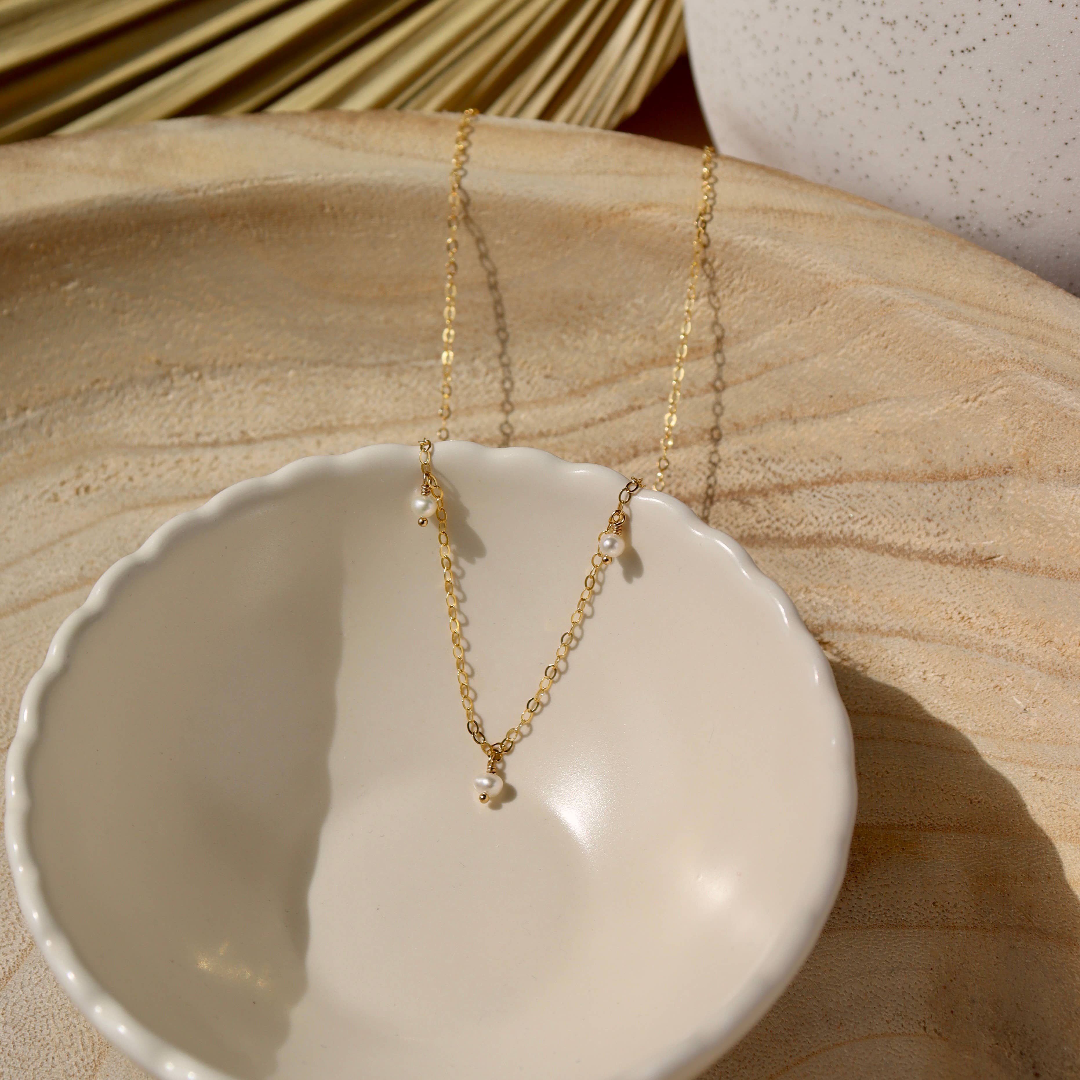 Delicate gold chain pearl necklace displayed in a white ceramic dish on a wooden surface