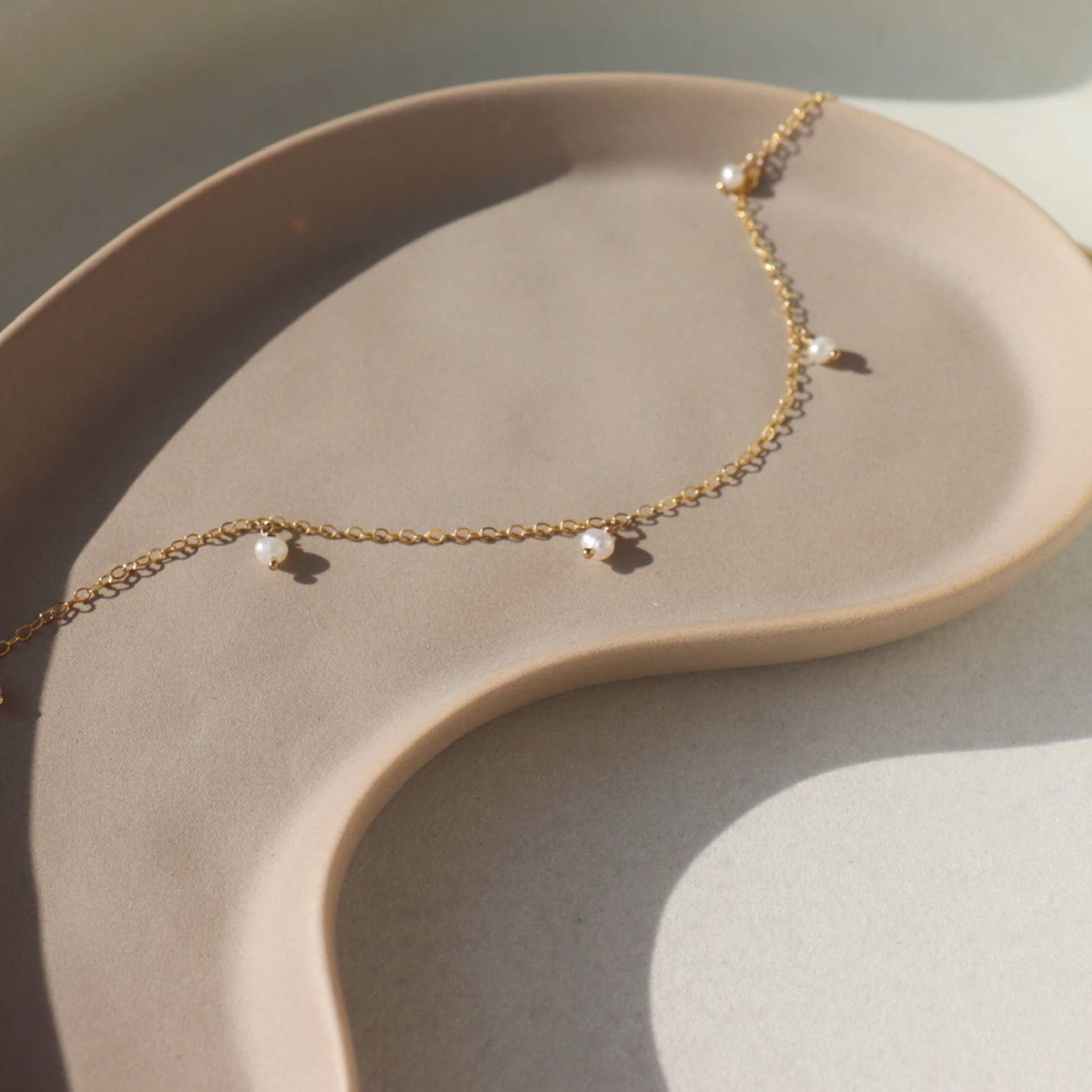 Delicate gold chain pearl necklace displayed on a soft beige dish