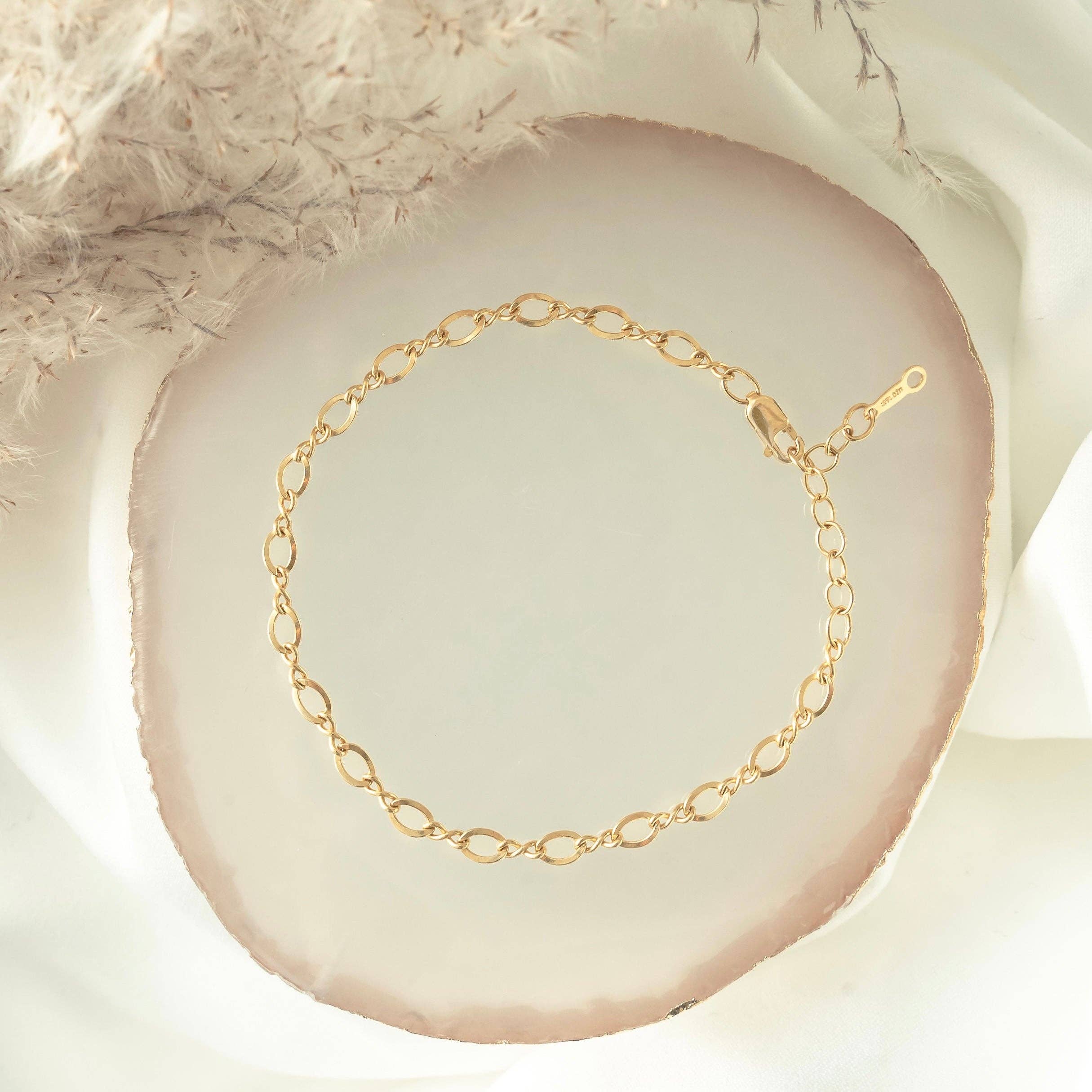 Elegant gold chain bracelet with oval links displayed on a white and gold-rimmed dish, styled with soft, neutral fabric for a delicate and refined look