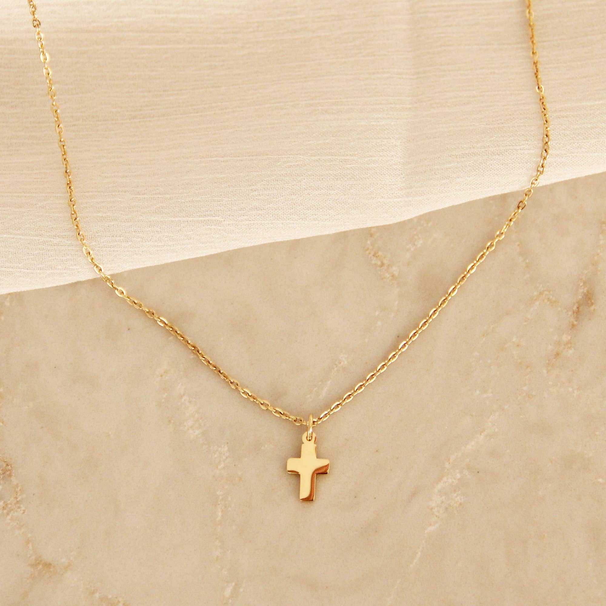 Gold cross pendant necklace displayed on a beige marble surface with a soft fabric accent