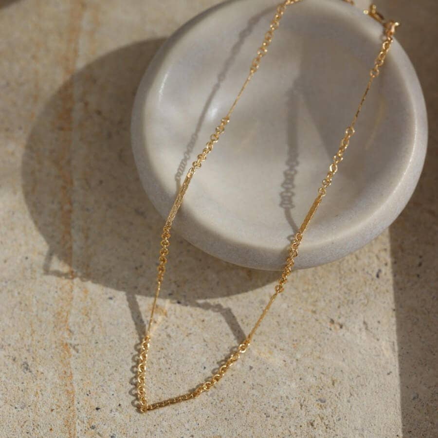 Gold bracelet chain, elegantly draped over a beige ceramic dish, casting shadows in the sunlight.