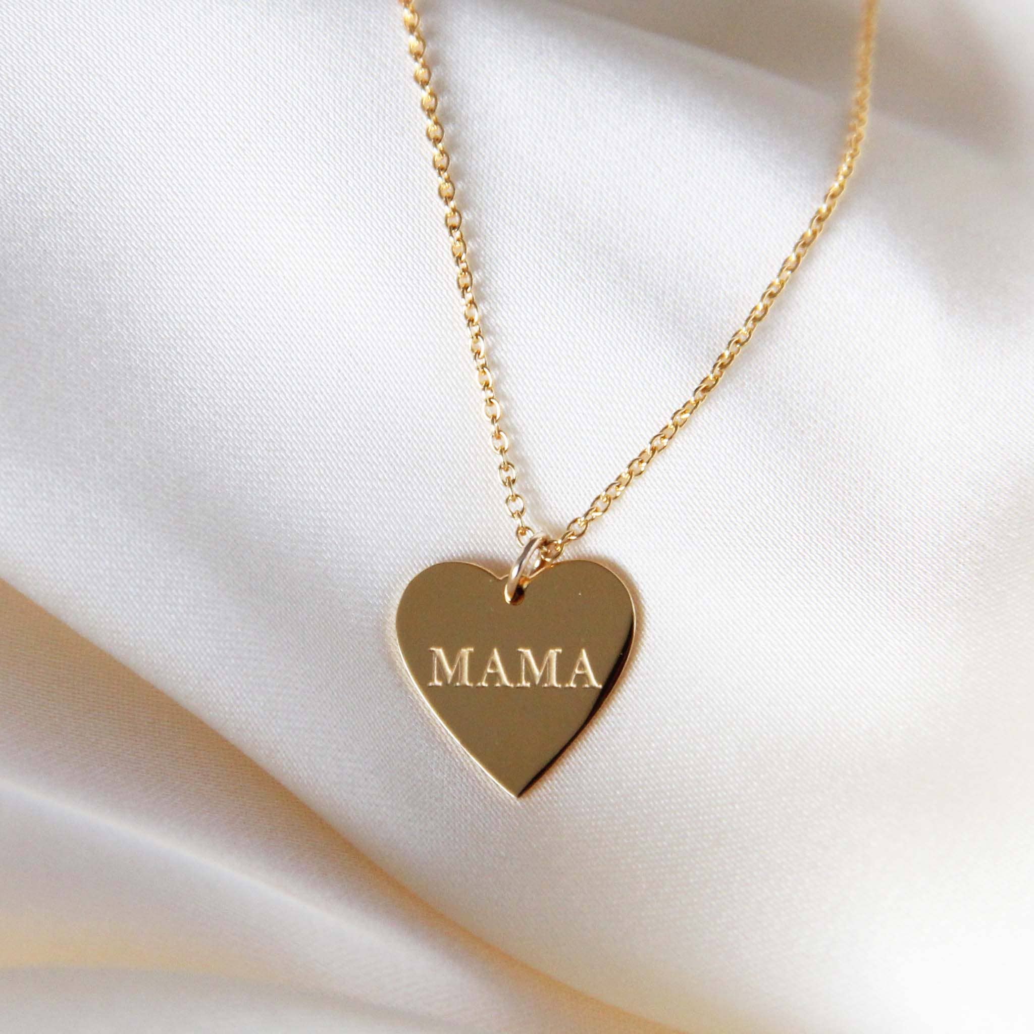 Gold heart-shaped 'Mama' necklace resting on a satin fabric, featuring an engraved 'MAMA' inscription on a delicate chain