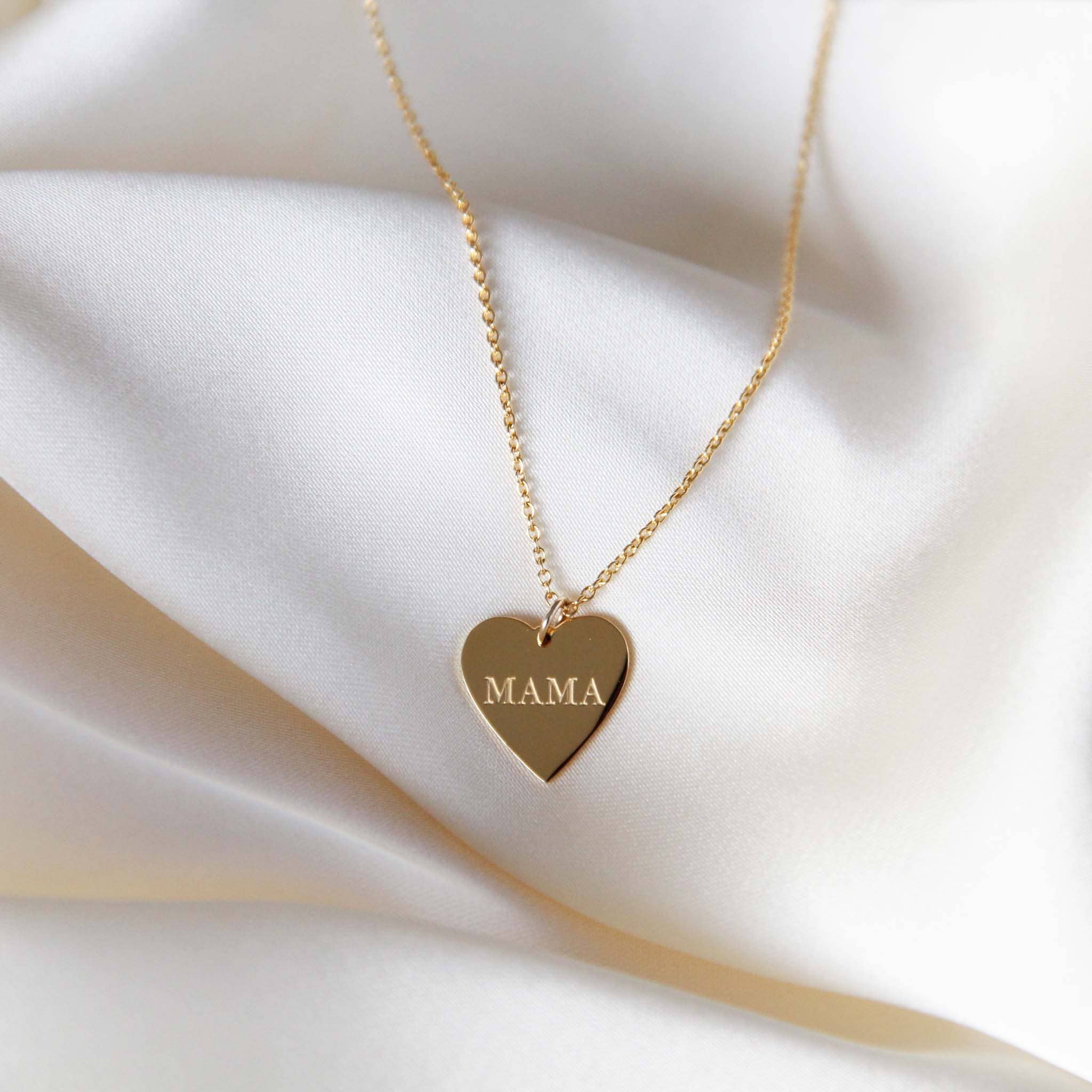 Gold heart-shaped 'Mama' necklace displayed on smooth satin fabric with an engraved 'MAMA' inscription on the pendant