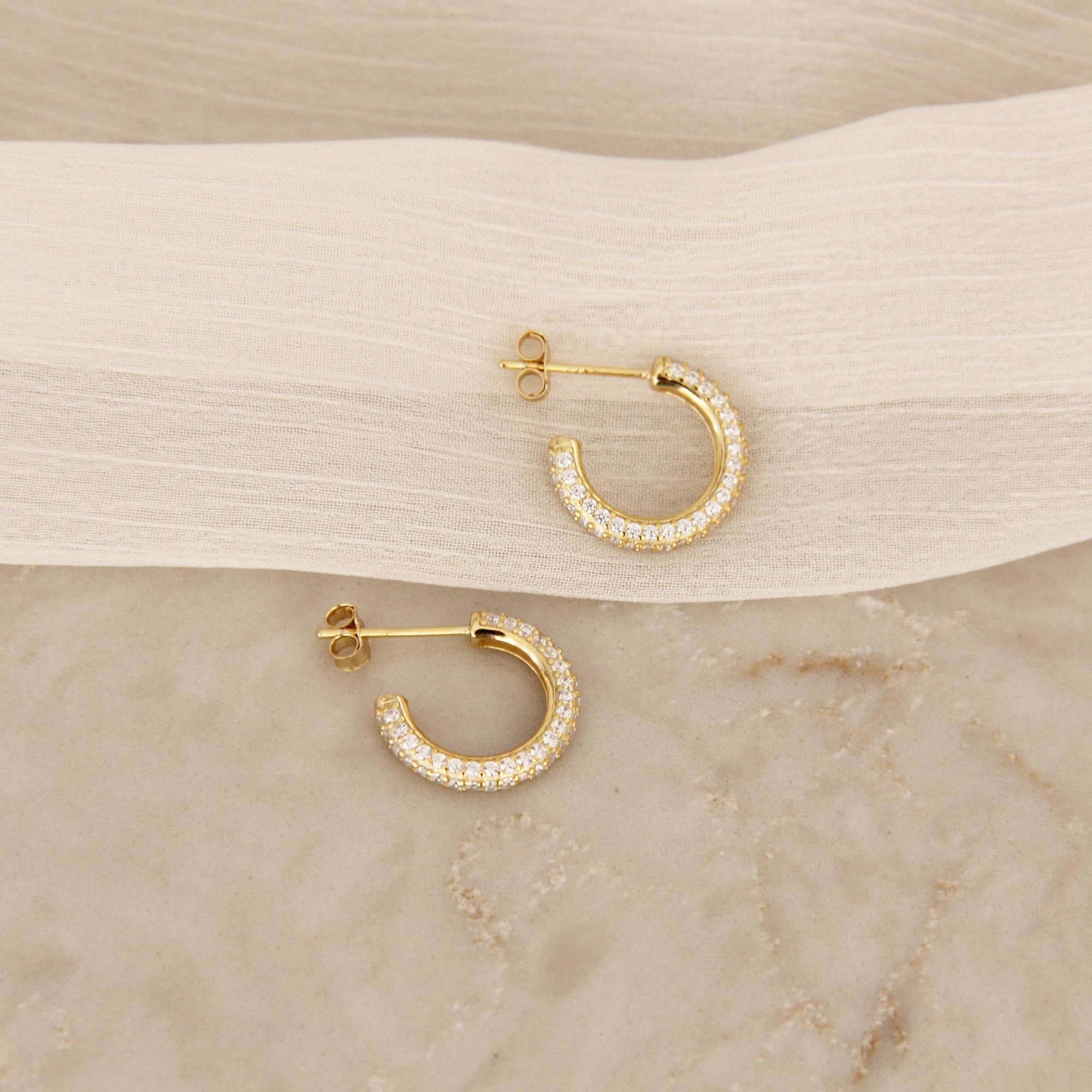 Gold cubic zirconia pavé hoop earrings displayed flat on a marble surface with a fabric drape, showcasing a different angle