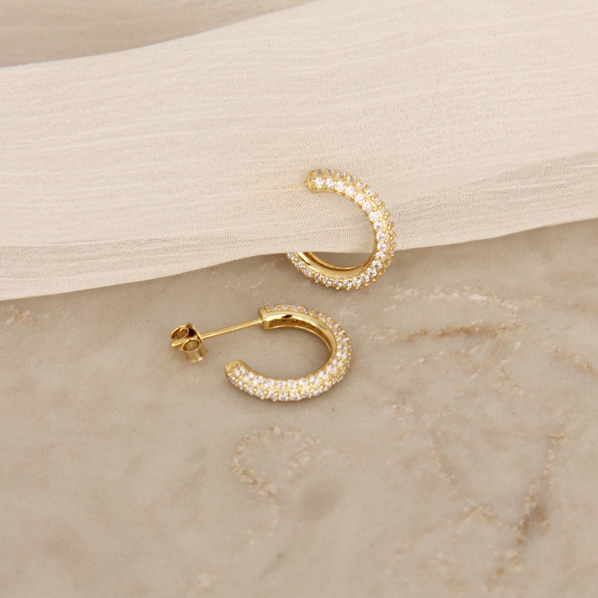 Gold pavé hoop earrings with cubic zirconia stones displayed at an angle on a marble surface with fabric drape