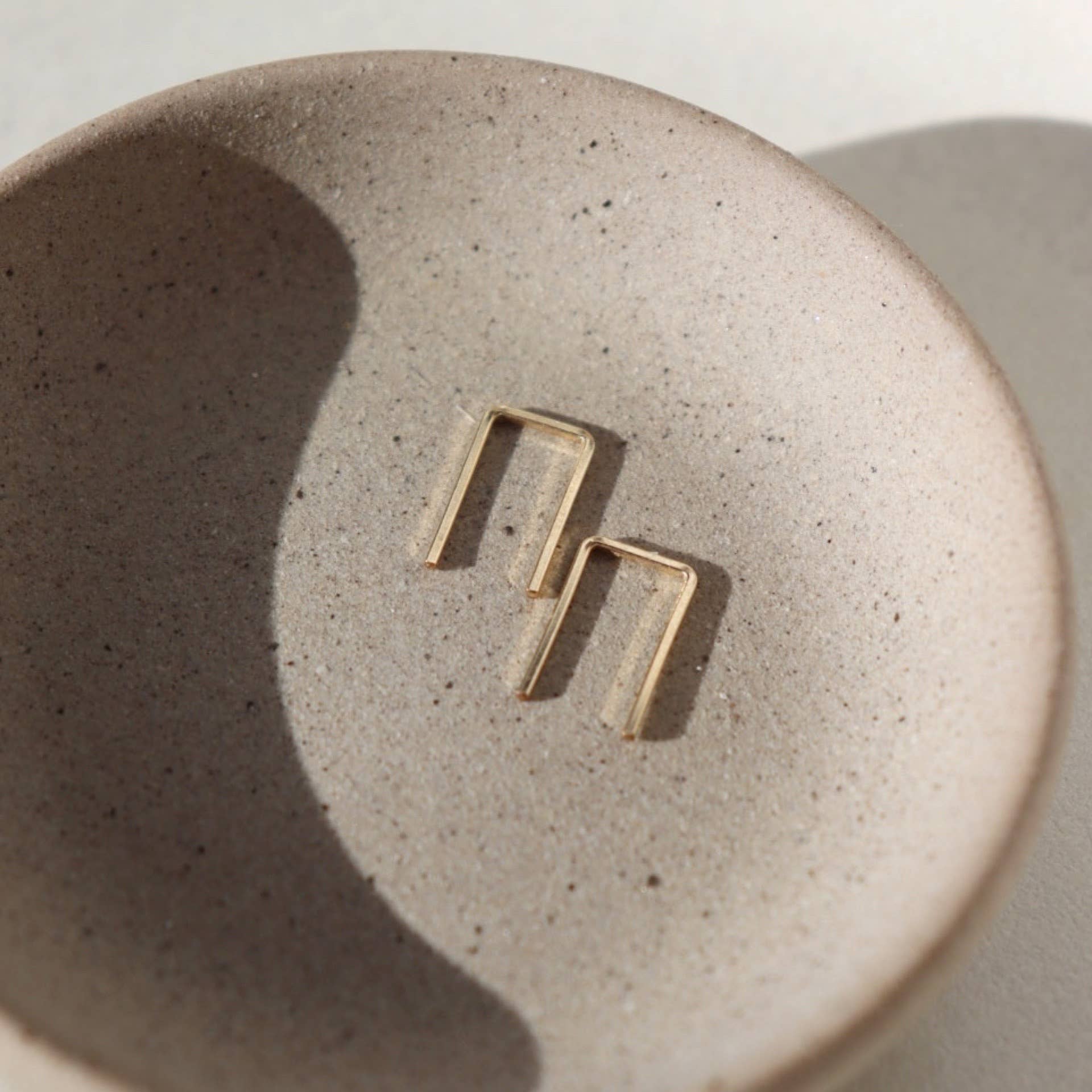 Minimalist gold rectangular staple earrings displayed in a textured neutral stone dish, illuminated by soft natural lighting.