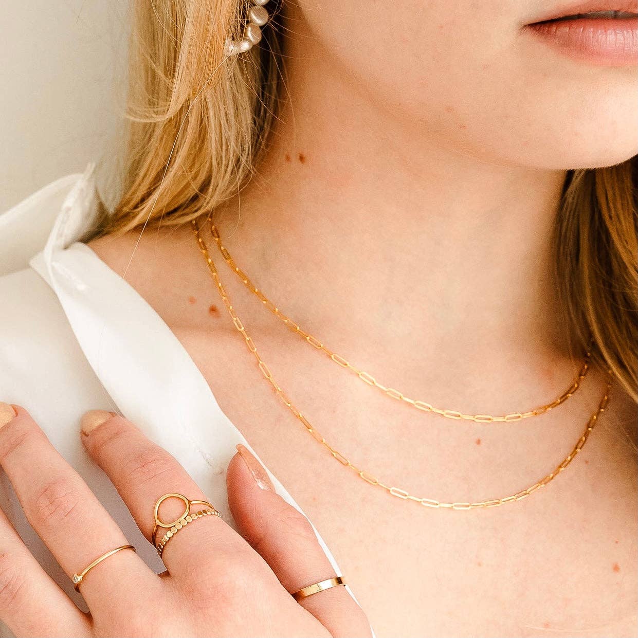 Close-up of a model wearing a layered gold chain necklace with elongated links, styled with pearl earrings and multiple rings for a chic, minimalist look.