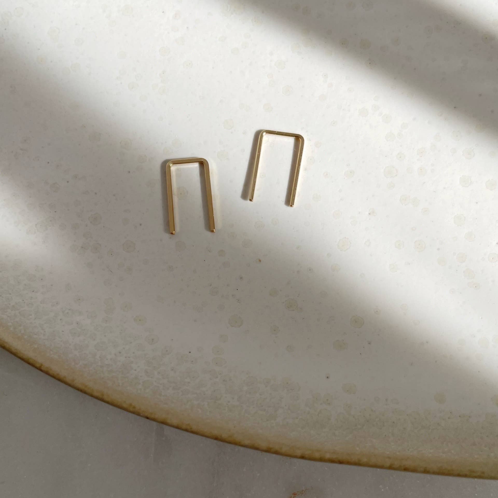 Minimalist gold staple earrings displayed on a textured surface with light shadow play, showing a modern and clean design.